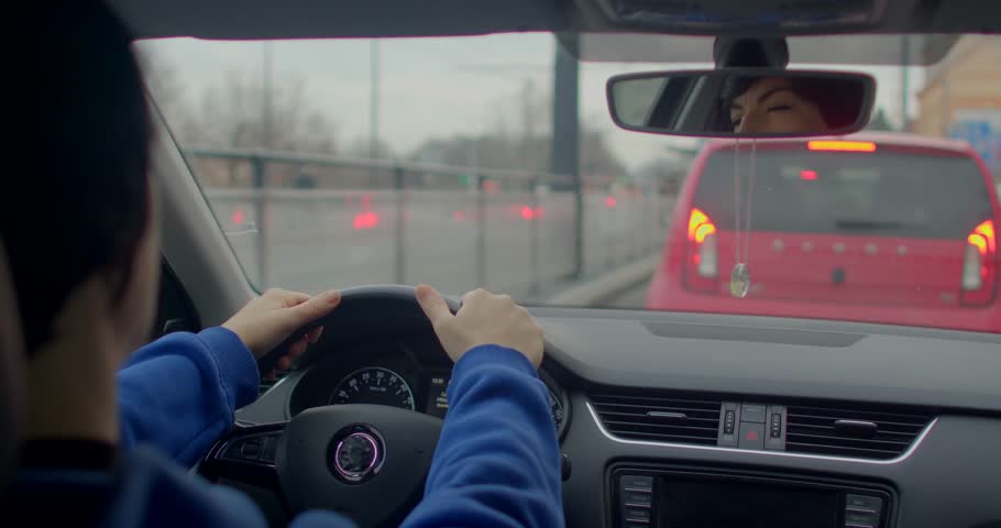 A driver sees a vibrant red car in front, surrounded by a cloudy urban backdrop and city bustle