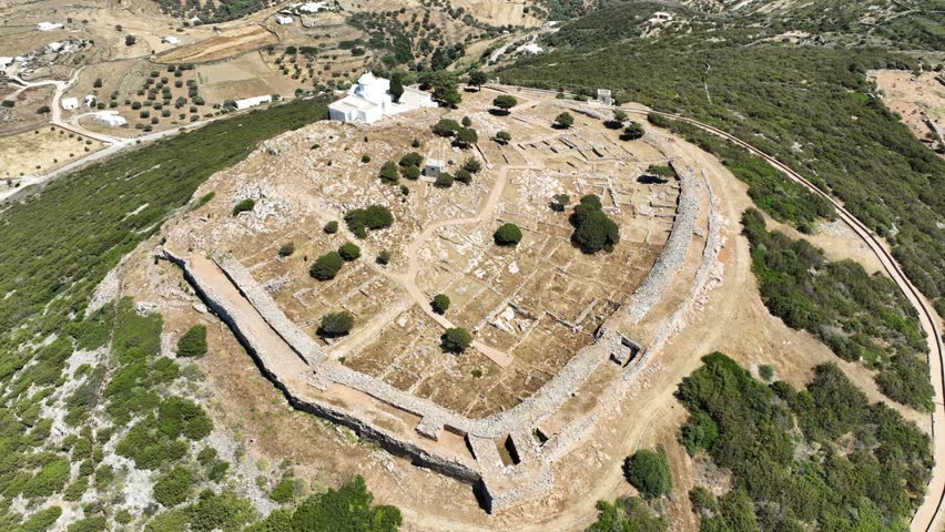 Aerial drone cinematic video of archaeological site of the Mycenaean Citadel and chapel of Agios Andreas built on top of hill overlooking the Aegean sea, Sifnos island, Cyclades, Greece