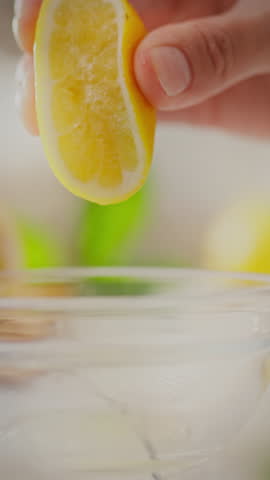 A hand is squeezing a slice of lemon, releasing juice into a clear bowl nearby. The vibrant yellow fruit contrasts with the fresh greens in the background, indicating a cooking or mixing activity