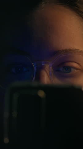 A young adult focuses intently on their smartphone screen, illuminated in a dark room. The soft light creates a captivating atmosphere, highlighting their glasses and facial features