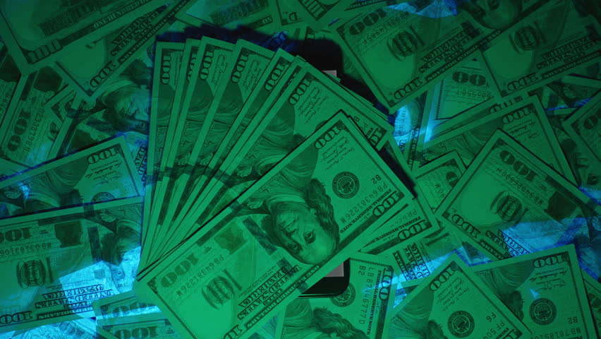 Overhead Shot of a Smartphone Displaying an Incoming Call Labeled "SCAM," Surrounded by US Hundred-Dollar Bills with a Hand Holding a Wad of Money in the Foreground, Representing How Scammers Exploit 