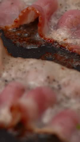 Close up of pieces bacon is frying in hot frying pan in sizzling oil and is sprinkled with spices and seasonings. Cooking meat. Vertical shot