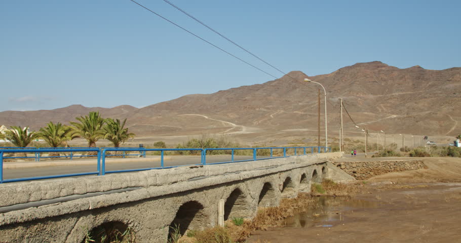 Historic Stone Bridge with Blue Railings in Gran Tarajal, Fuerteventura Against Desert Mountains. Ancient Aqueduct-Style Architecture with Palm Trees and Mountain Road in Canary Islands Landscape