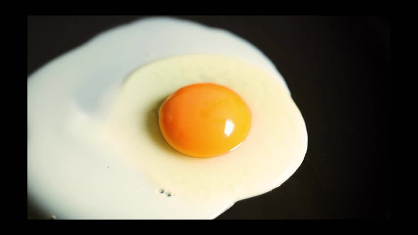 An egg is cracked into a hot black pan, and as it cooks, the egg white turns white and firms up.