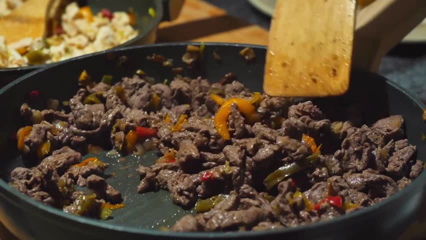 A person is julienning meat and preparing fajitas, mixing it in a pan with red peppers using a wooden spoon. The vibrant ingredients cook together, creating a delicious, sizzling dish.