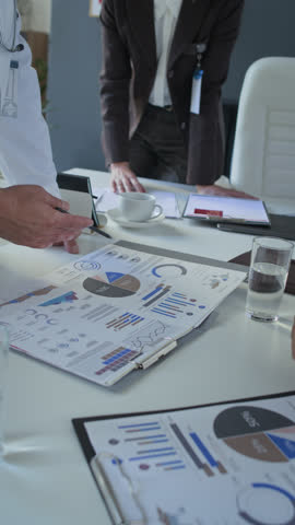 Vertical close-up shot of unrecognizable doctor pointing at charts and statistics on paper during meeting with colleagues in medical office