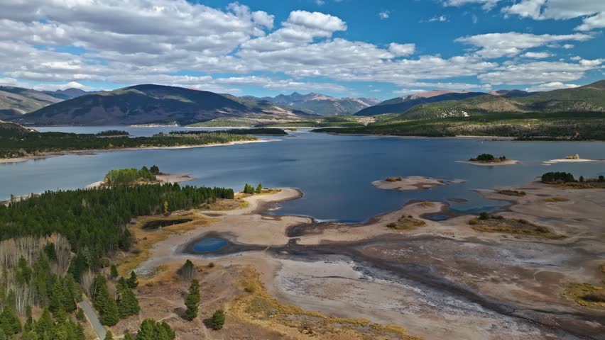 Aerial of lake shore with winding inlets, surrounded by mountains under a cloudy sky, drone pullback