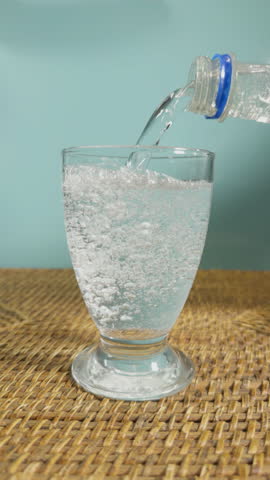 Vertical video - Closeup normal speed then slow motion of clear sparkling water being poured from a bottle into a drinking glass.