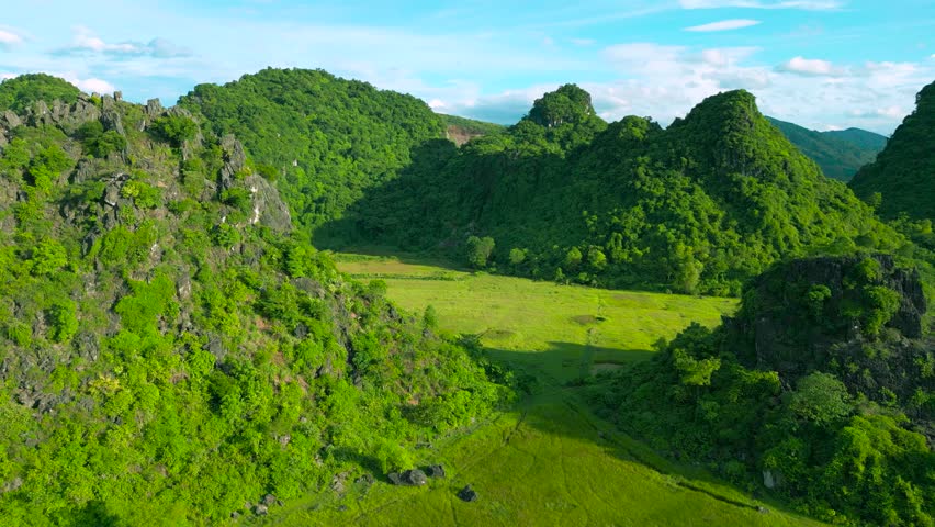 Lush tropical mountains envelop Phong Nha, Ke Bang National Park in Vietnam, showcasing a vibrant green landscape, rocky formations, and open fields under a clear blue sky.