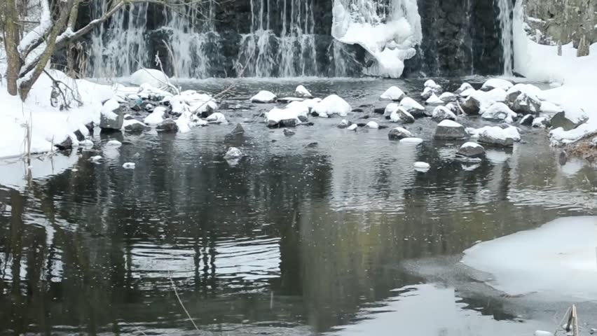 Frozen river with a waterfall. The water is still and the rocks are covered in snow. The scene is peaceful and serene