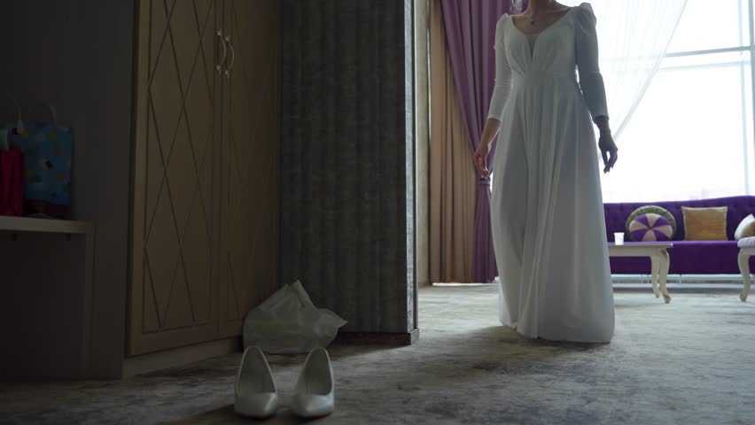 Bride in white wedding dress walks through the room with window and puts her feet in female shoes. Concept of festive or wedding event, traditional elements of classic women