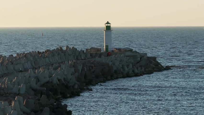A lighthouse with a green top stands on a concrete block breakwater, extending into the sea. Sailboats dot the ocean under a clear sky.