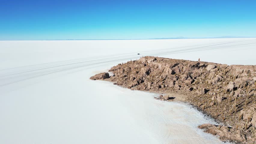 A drone captures a 4WD vehicle driving across the vast Salar de Uyuni salt flats, passing rocky mountain terrain during the day in Bolivia