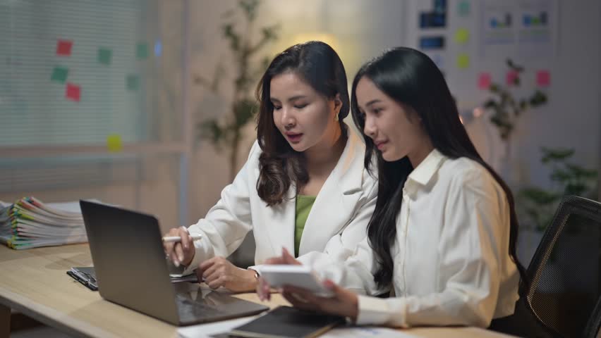 Two women are sitting at a desk with a laptop and a tablet. They are looking at the laptop screen and discussing something