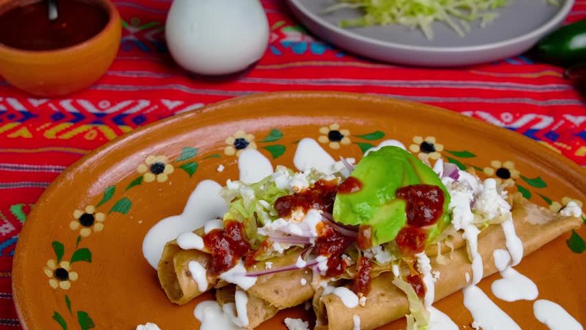 Taquitos garnished with lettuce, mexican cream, cheese, onion and avocado, served on a clay plate, mexican food