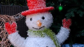 A snowman with a red hat and green scarf is standing in front of a Christmas tree. The snowman is smiling and holding a red glove in its right hand. The scene conveys a festive and joyful mood - Powered by Shutterstock - Get 15% off with code: PIKWIZARD15