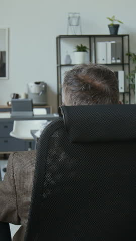 Vertical portrait shot of middle-aged businessman in formal suit sitting on office chair, putting on blue clown wig and red nose, then turning to camera and posing during workday