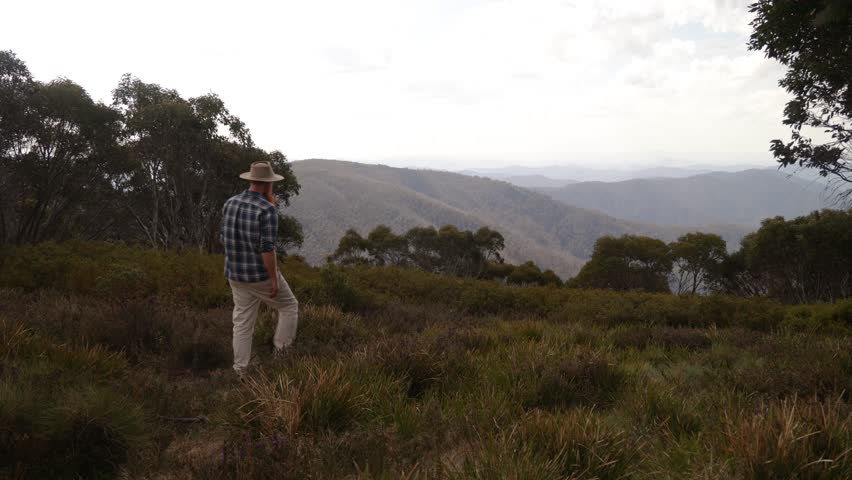 Australian bushman walks towards the edge of a mountain range lookout in the Australian wilderness.