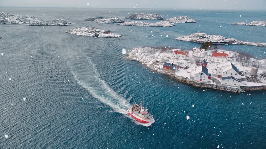 Aerial view of floating fishing boat in blue sea in snowfall. Henningsvaer, Lofoten islands, Norway in winter. Top drone view of boat, mountains in snow, sea, cloudy sky, town, rorbuer houses. Travel