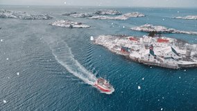 Aerial view of floating fishing boat in blue sea in snowfall. Henningsvaer, Lofoten islands, Norway in winter. Top drone view of boat, mountains in snow, sea, cloudy sky, town, rorbuer houses. Travel - Powered by Shutterstock - Get 15% off with code: PIKWIZARD15