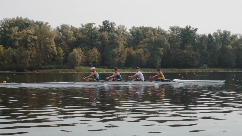 Rowing team training. Side view of 4 young caucasian male rowers, during a rowing practice, athlete sitting in a boat in the river Dnipro, rows through a calm water in autumn. 4k footage. City area in