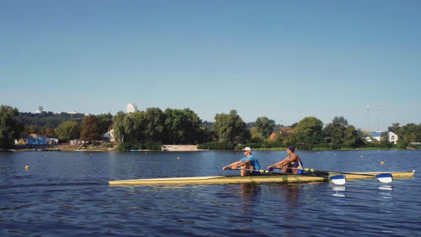 Rowing team training. Side view of 2 young caucasian male rowers, during a rowing practice, athlete sitting in a boat in the river Dnipro, rows through a calm water sunny day, autumn. 4k footage