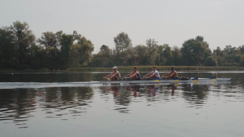 Rowing team training. Side view of 4 young caucasian male rowers, during a rowing practice, athlete sitting in a boat in the river Dnipro, rows through a calm water in autumn. 4k footage. City area in