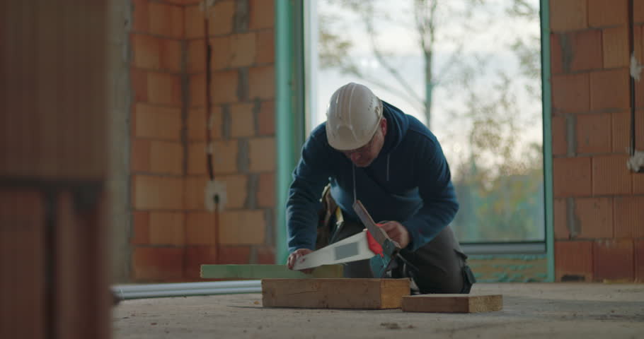 Construction worker kneeling and sawing wood with handsaw on indoor construction site, focused on precise work, surrounded by unfinished walls, building project in progress
