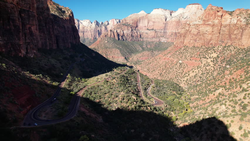 Aerial view of a picturesque valley featuring winding roads, rugged cliffs, and vibrant desert vegetation under a clear blue sky.