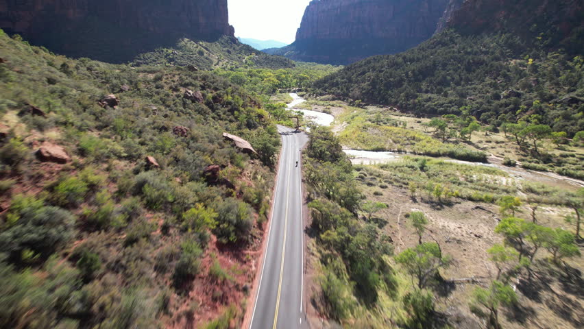 A dynamic aerial view following a road through a serene valley, seamlessly transitioning to track a winding river framed by lush greenery and dramatic cliffs in Zion National Park.