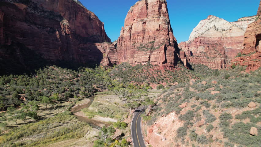 A stunning aerial shot flying along a road winding through Zion National Park, framed by towering red sandstone cliffs and lush greenery under a vibrant blue sky.