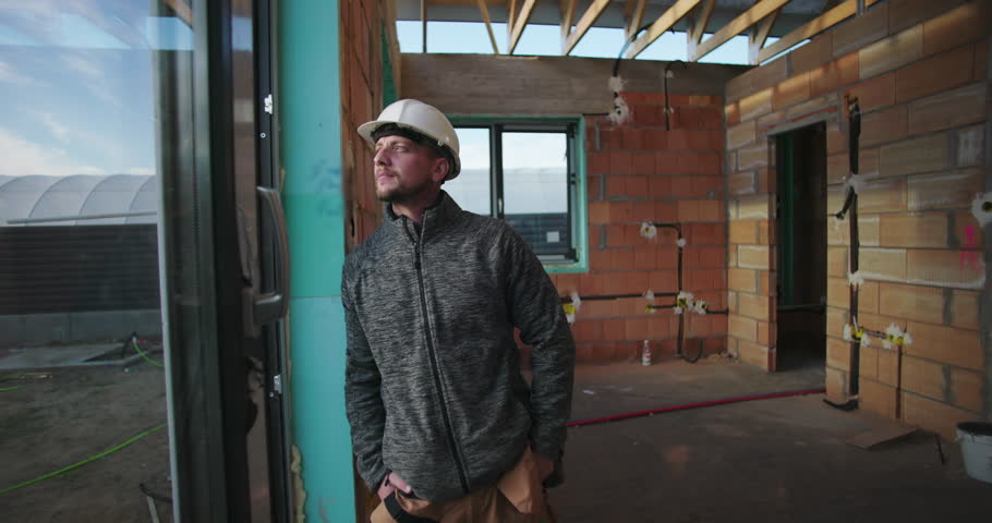 Young construction worker gazing outside, hands in pockets, hard hat on, inside partially constructed building, with unfinished brick walls and window frame in background
