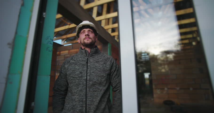 Young construction worker with arms crossed, wearing a hard hat, looking determined, standing by a glass wall with wood framework in the background on a construction site