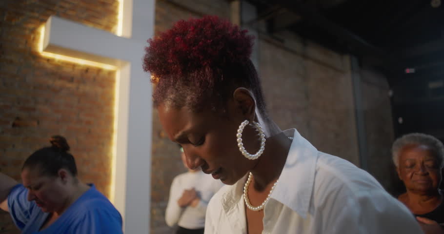Elderly woman in church placing hands on chest, eyes closed in prayer, illuminated cross and brick wall in background, community worship and spiritual connection