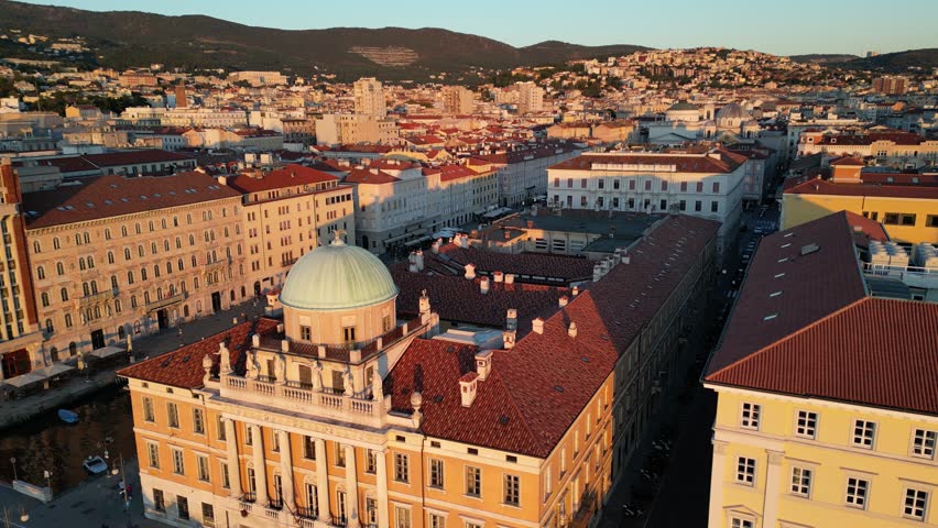aerial view of downtown trieste (historic buildings on Mediterranean coast canal, famous buildings) boats in marina, cars driving road (sunset golden hour) red roof piazza palazzo footage drone italy