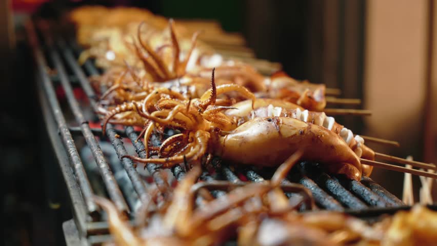 Close-up of grilled seafood skewers on stick. Man hand cutting with scissors and flip. Yaowarat Road in Chinatown - popular night market for try street food. Bangkok, Thailand