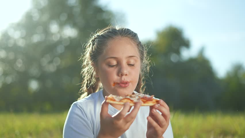 Smiling child enjoying pizza at picnic in nature. Happy girl eating pizza outdoors during family picnic. Sunny day picnic with pizza brings joy to child. Fun outdoor pizza picnic in nature with child.