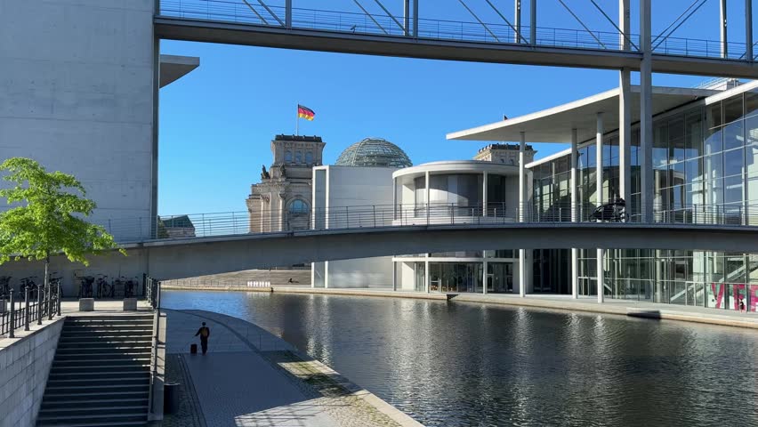 Modern architecture in Berlin featuring the Reichstag with its glass dome in the background, a cyclist on a bridge, and a reflective water canal in the foreground under a bright blue sky