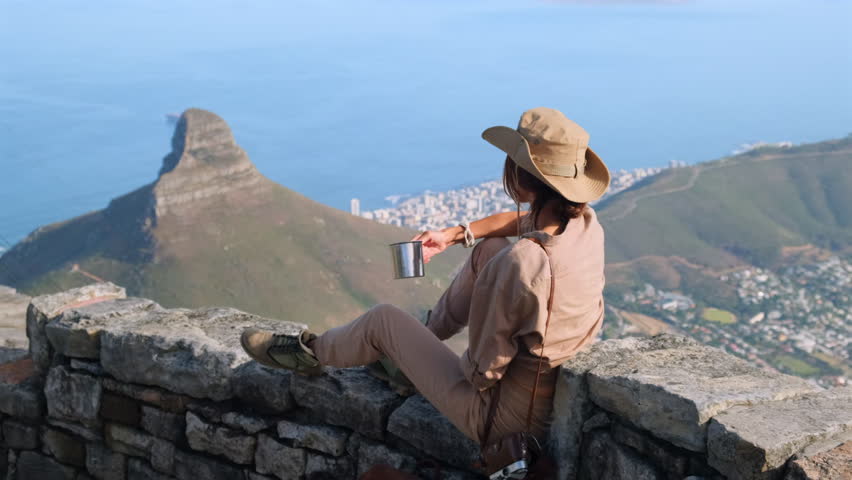 girl hiker in boots drinking a hot drink from a mug sitting on a mountain peak relaxing after hiking and taking a break. A hiker wearing a sun hat and sunglasses, looking out over a scenic vista
