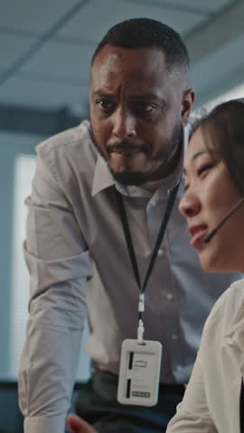 Call center office: African American technical support specialist helps Asian colleague in headset. Two multiethnic hotline operators working in online customer assistance service, using computer.