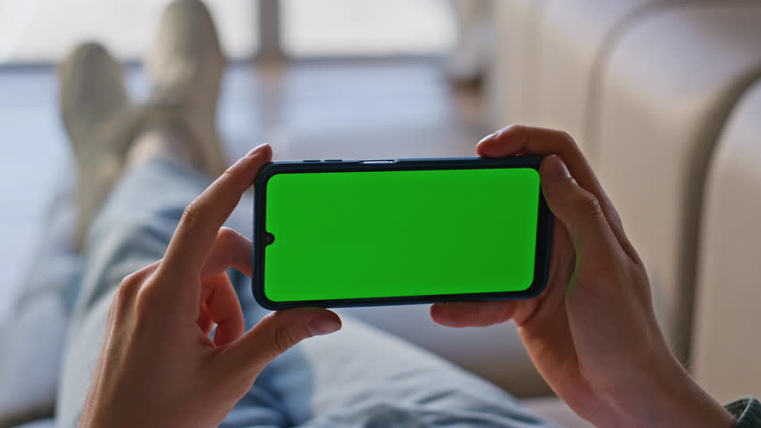 Closeup hands holding chromakey mobile phone at home. Unrecognized man watching greenscreen cellphone resting couch. Unknown guy using mockup smartphone lying sofa at weekend room. Technology concept