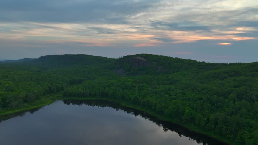 A breathtaking view of Lake of the Clouds at sunset during July, captured by drone footage in the Porcupine Mountains, Upper Peninsula, Michigan. The tranquil lake reflects the serene sky.