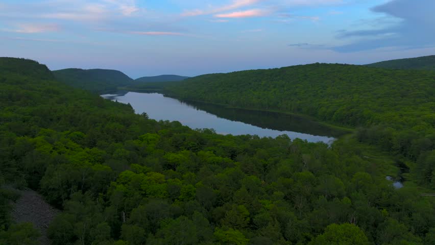 A breathtaking view of Lake of the Clouds at sunset during July, captured by drone footage in the Porcupine Mountains, Upper Peninsula, Michigan. The tranquil lake reflects the serene sky.
