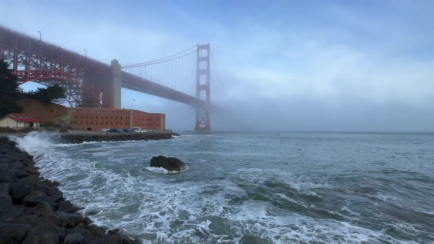 Foggy mist Golden Gate Bridge surf wave crash on rocks San Fransisco California Fort Point National Historic site vista point welcome center cloudy morning Pacific Ocean sea warming hut static shot