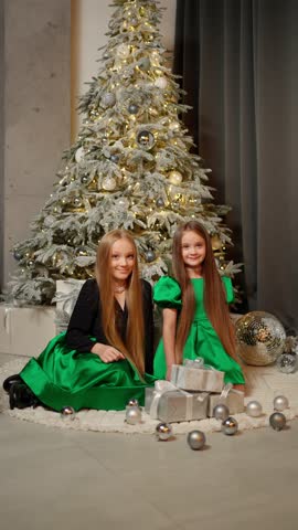 Two girls in festive clothes are sitting near Christmas tree. Concept of celebrating new year.