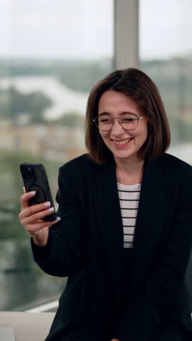 Young lady with dark short hair looks at her smartphone. Woman in glasses laughs cheerfully using her phone. Vertical video.