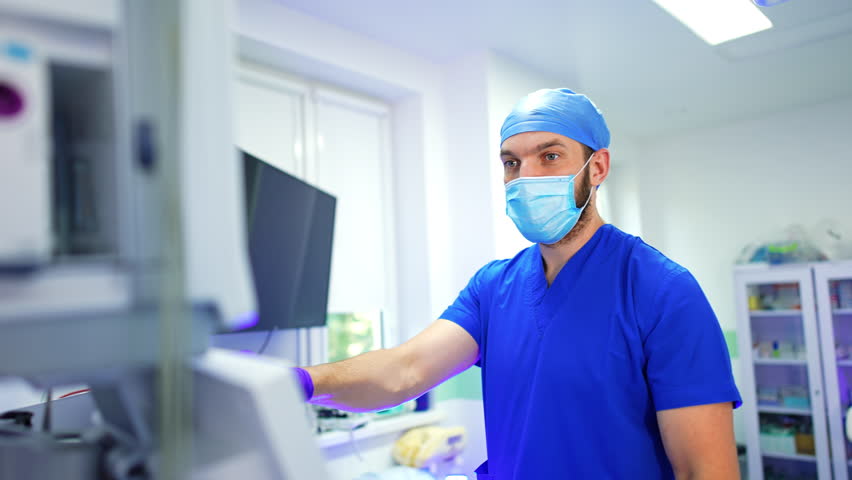 Mid-aged medic in robe, mask, cap and gloves presses the keys on the medical equipment. Anesthesiologist working at operation in modern surgery room.