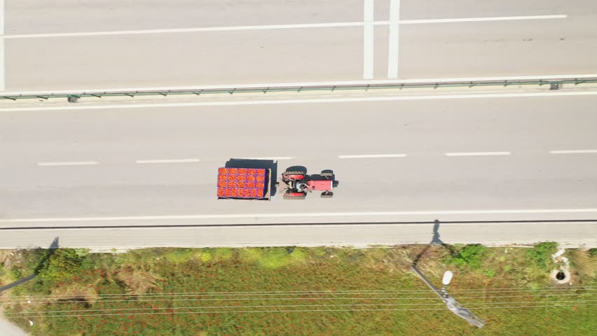 Aerial view of a red tractor carrying a full trailer driving on the road next to agricultural fields during harvest season