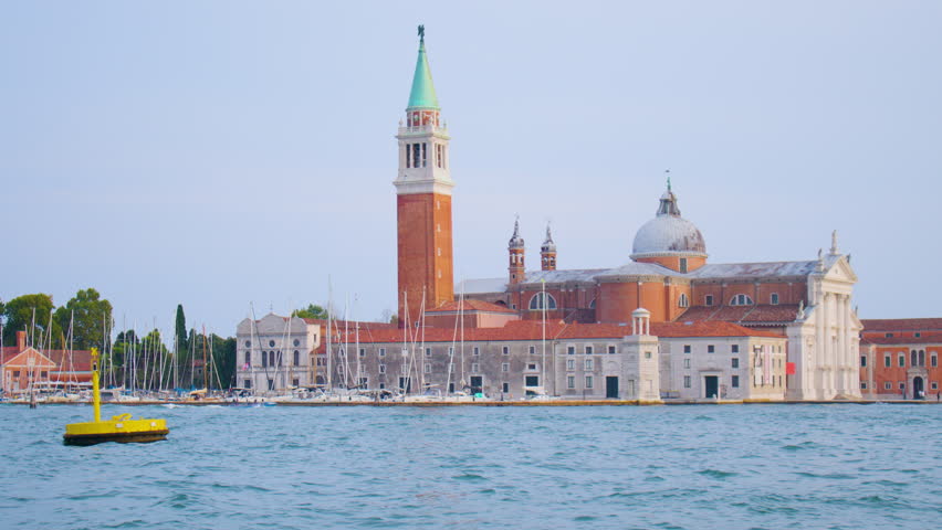 A view of San Giorgio Maggiore island and church from water, with rows of yachts and boats in the foreground. Sailing near the historical church on an island, surrounded with multiple docked boats.