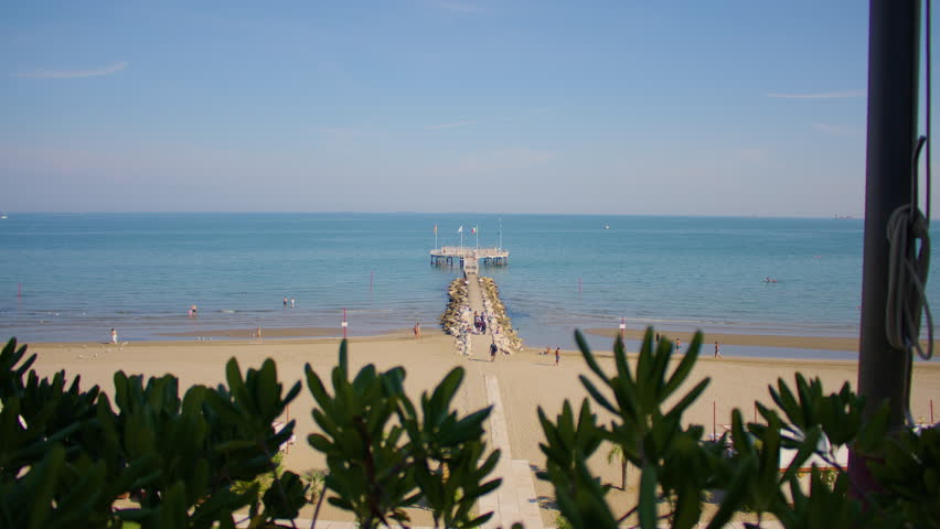 Wide shot of an empty sandy beach, with calm sea, long pier and greenery in the foreground. A view of beach and pier near Venice lagoon on a bright sunny day.
